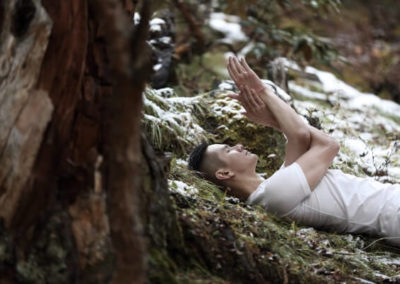 Chris Su lying on the ground in a forest wearing all white doing eagle pose with his arms