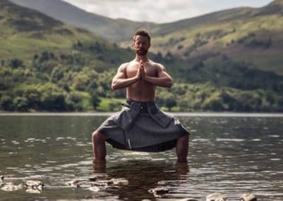 Finlay Wilson topless in Scottish tilt standing in low water with green hills in the background